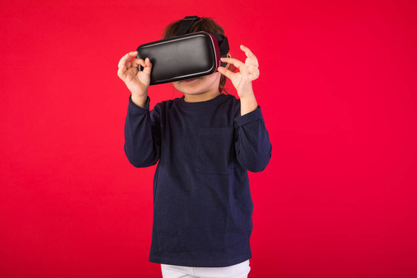 Klein meisje in virtual reality bril verbaasd en aanraken met haar handen virtueel, op rode achtergrond. Virtuele, augmented reality, technologie, games en jeugdconcept. - Foto, afbeelding