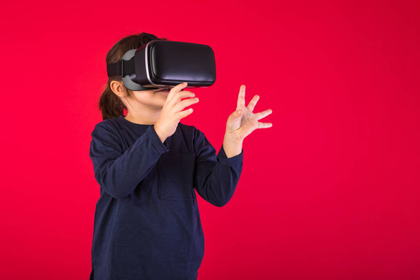 Side view of little girl wearing virtual reality goggles amazed and touching with her hands virtually, on red background. Virtual, augmented reality, technology, games and childhood concept. - Foto, afbeelding