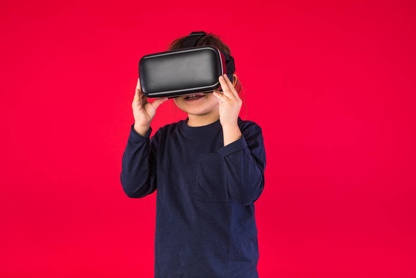 Front view of little girl wearing virtual reality glasses amazed and touching with her hands virtually, on red background. Virtual, augmented reality, technology, games and childhood concept. - Photo, Image