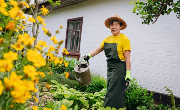 Mujer jardinera mayor en un sombrero trabajando en su patio con herramientas de trabajo. El concepto de jardinería, cultivo y cuidado de flores y plantas. - Foto, imagen