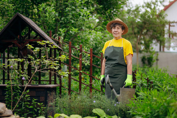 Starší zahradnice v klobouku, pracující na jejím dvorku s pracovním nářadím. Koncept zahradničení, pěstování a péče o květiny a rostliny. - Fotografie, Obrázek