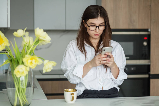 Brunette in eyeglasses. Beautiful vase with yellow flowers. Cozy kitchen. Modern interior in shades of grey. Brunette girl with healthy lifestyle relaxes at home in morning. - Fotografie, Obrázek