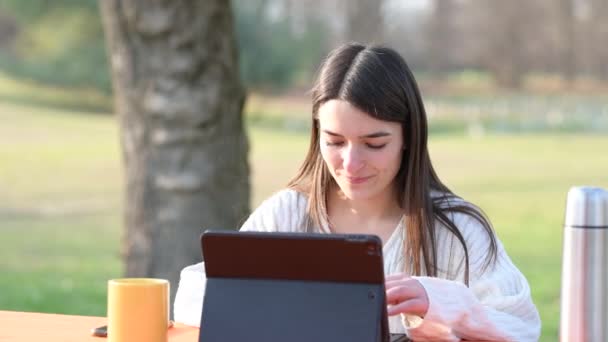 Portret van een jonge mooie vrouw die op afstand werkt in een park, drinkt uit een groene beker en typt op haar laptop toetsenbord. De wind beweegt haar haar licht. Op de achtergrond wazig mensen. - Video