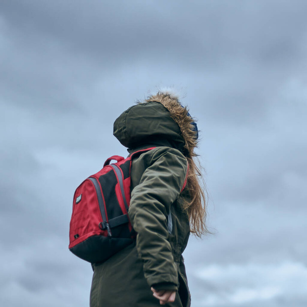Girl with long hair, a red backpack and a green coat jacket. Beautiful nature background. - Photo, Image