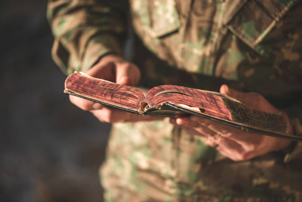 Στρατιώτης ντυμένος με στολή καμουφλάζ κρατώντας μια Βίβλο στο χέρι του. Στρατιώτης που διαβάζει και διαλογίζεται στο λόγο του Θεού - Φωτογραφία, εικόνα