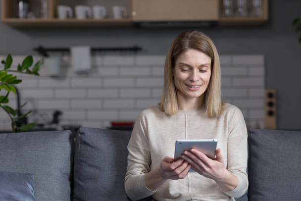 Happy woman relaxing at home, using tablet computer sitting on sofa in kitchen - Photo, Image