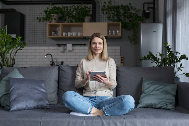 Woman sitting on sofa at home, using tablet computer, reading e-books and online learning - Foto, Bild