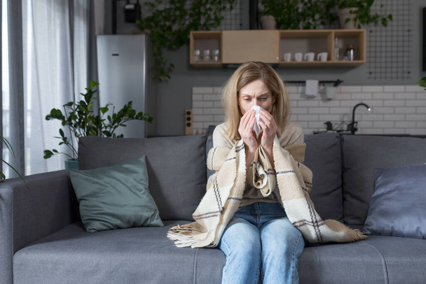 The sick woman is sitting on the couch at home, has a cough and fever, cold and runny nose - Foto, Imagen