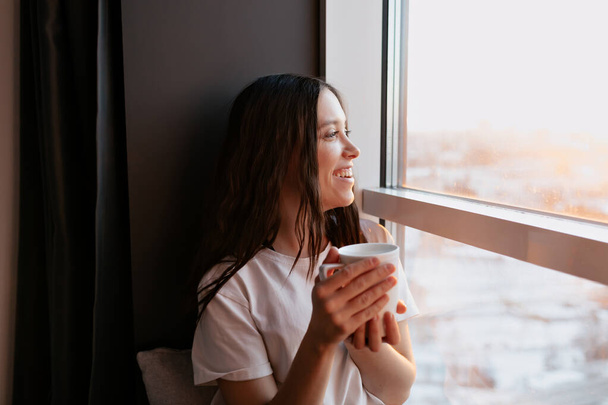 Smiling happy incredible girl with dark hair wearing white t-shirt is sitting near the window in sunshine and drinking morning coffee. Stylish girl at home in the morning. High quality photo - Photo, Image