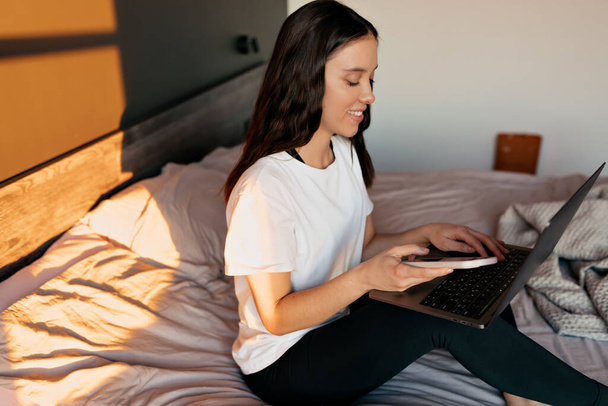Mooie schattige vrouw met donker haar draagt thuis outfit typt op laptop in het bed in de ochtend zonlicht. De vrouw werkt vanuit huis. . Hoge kwaliteit foto - Foto, afbeelding