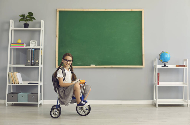 Happy little student girl riding bicycle inside school classroom with copy space chalkboard - Foto, Imagen