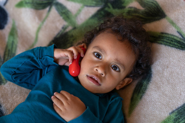 Latin American boy (2) holds a toy headset to his ear while playing on the phone. Children's technology concept - Foto, imagen