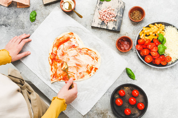 白人の10代の少女の手は、テーブルの上の食材でバレンタインの日のためのハート型のピザ生地にソースのスプーンをスミア、トップビューのクローズアップ。バレンタインデーのピザ作りのコンセプト. - 写真・画像