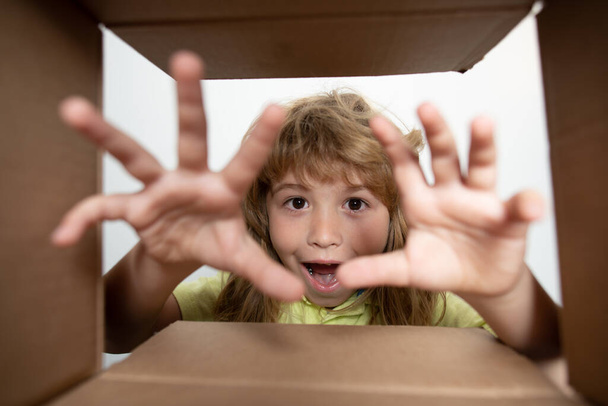 Child opens carton box and pulling out gift from it, take by hand gift. Small boy looking in parcel box and happy to receiving a surprise. Shocked kids, amazed kids emotions, wow. - Valokuva, kuva