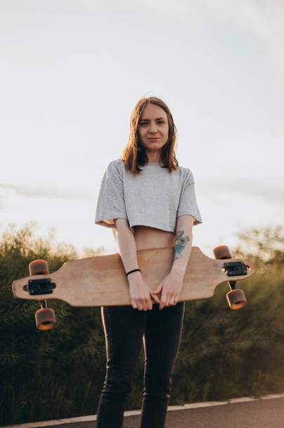 Front view , low angle .Funny young girl in jeans and top stands with big longboard. Green nature background - Foto, imagen