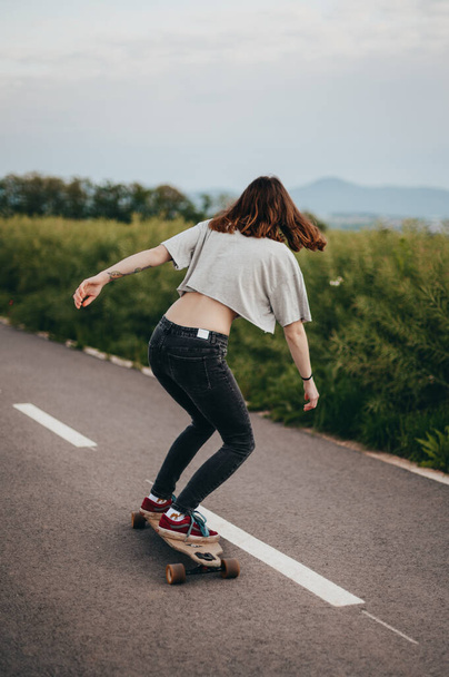 Vue de l'arrière, skateboarder féminin à cheval longue planche sur la piste cyclable, prairie verte en arrière-plan - Photo, image