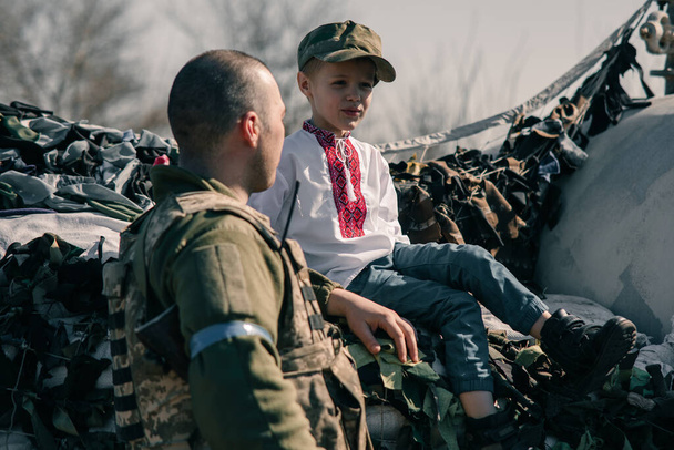Child boy in embroidered shirt sits on sandbags in checkpoint near Ukrainian territorial defense warrior. Concept of russian military invasion in Ukraine. War in Ukraine and Europe. Children and war. - Photo, Image