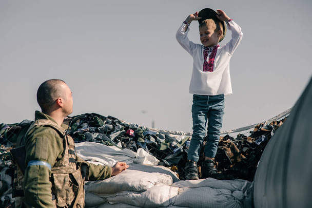 刺繍シャツの子供の男の子は、ウクライナの領土防衛戦士の近くのチェックポイントのサンドバッグに立っています。ウクライナにおけるロシアの軍事侵攻の概念。ウクライナとヨーロッパでの戦争。子供と戦争 - 写真・画像