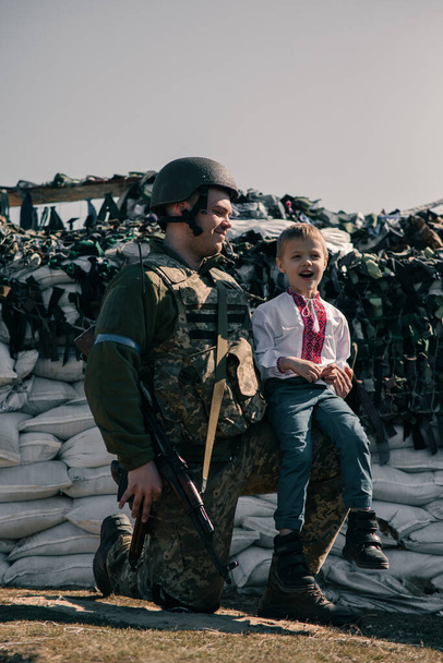 刺繍シャツの子供の男の子は、サンドバッグの背景に対するチェックポイントにウクライナの領土防衛戦士の近くに座っている。ウクライナにおけるロシアの軍事侵攻の概念。ウクライナとヨーロッパでの戦争。子供と戦争. - 写真・画像