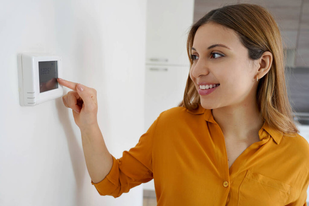 Πορτρέτο της χαμογελαστής γυναίκας μειώνει τη θερμοκρασία στον ψηφιακό θερμοστάτη στο σπίτι. Εξοικονόμηση ενέργειας, αποδοτική και έξυπνη τεχνολογία. - Φωτογραφία, εικόνα