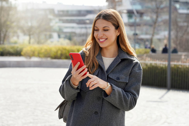 Mittlere Aufnahme einer jungen lächelnden Frau, die an sonnigen Tagen ihr Handy auf der Straße benutzt - Foto, Bild