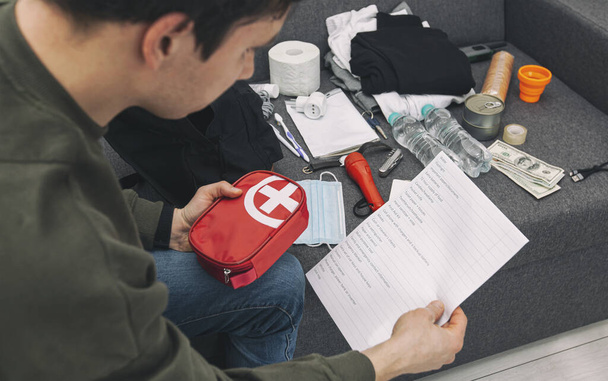 Young man packing the bag with documents, water,food, first aid kit and other items needed to survive - Photo, Image
