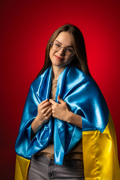 Νεαρή Ουκρανή γυναίκα με σημαία της Ουκρανίας μπλε και κίτρινο στέκεται πάνω από το κόκκινο φως φόντο - Φωτογραφία, εικόνα