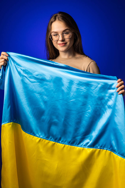 Jovem mulher com a bandeira nacional da Ucrânia em pé sobre fundo azul. - Foto, Imagem