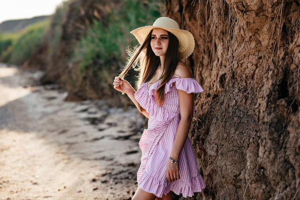 Young woman in dress and straw hat walk alone on empty sand beach at summer sunset on sea shoreand and smiling. - Foto, Imagem