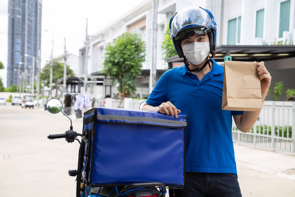 フェイスマスクを着用し、食品用紙袋を保持し、宅配ボックスの側に立っている若いアジアの男,食品エクスプレスサービスの概念を提供するオートバイ - 写真・画像