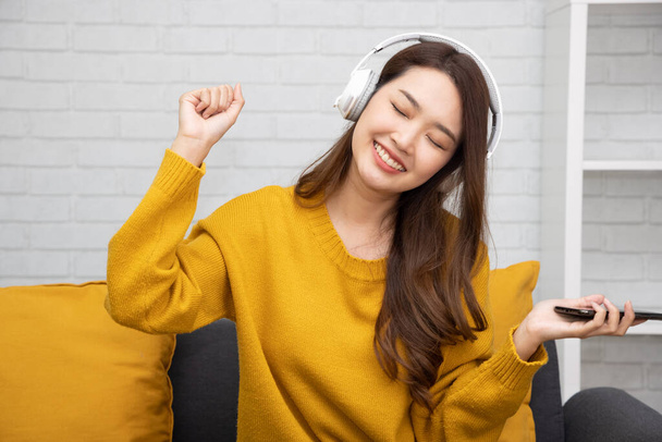 Ritratto di una giovane donna asiatica felice che ascolta musica con cuffie senza fili su un'applicazione musicale nel telefono cellulare e si siede sul divano nella stanza a casa - Foto, immagini