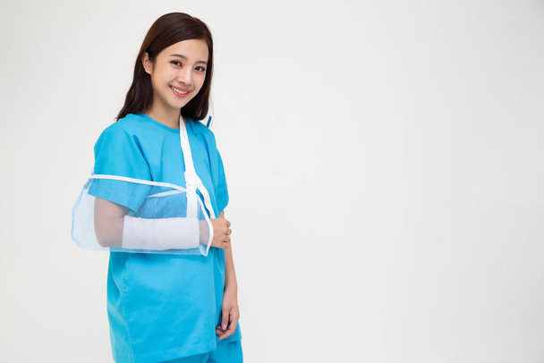 Hasta kıyafeti giyen genç Asyalı bir kadın beyaz arka planda izole edilmiş kırık bir kol yüzünden yumuşak bir atel taktı. - Fotoğraf, Görsel