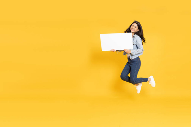Mladá asijská žena ukazuje a drží prázdné bílé billboard a skákání izolované na žlutém pozadí - Fotografie, Obrázek