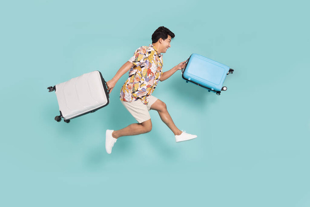 Homem asiático feliz viajante pulando e segurando mala isolada no fundo verde, turista tendo conceito de viagem de férias alegre, composição de corpo inteiro - Foto, Imagem