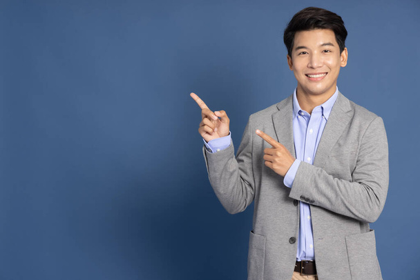 Giovane uomo d'affari asiatico sorridente e indicando spazio vuoto copia isolato su sfondo blu profondo - Foto, immagini