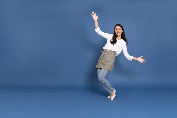 Young Asian woman housewife wearing kitchen apron cooking and dancing isolated on blue background - Fotoğraf, Görsel