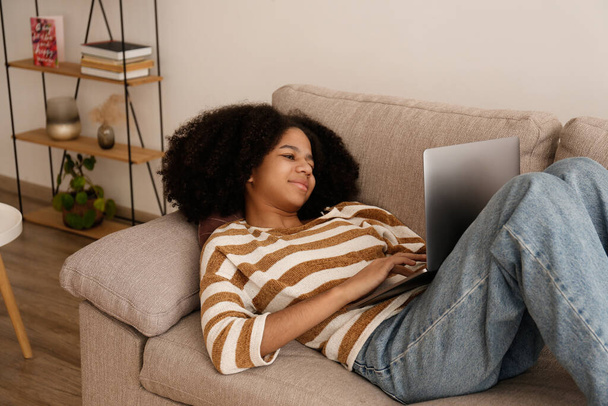 Black teenage girl with afro hairstyle lying on the couch, browsing the internet on her laptop. African american female wearing casual attire relaxing at home. Close up, copy space, background. - Фото, зображення