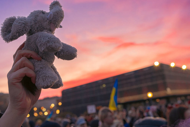 juguete de niño en la mano de la persona, elevado a la cima, en multitud de manifestantes contra la guerra en Ucrania. símbolo de la infancia y la libertad de los niños ucranianos. Nubes rojas en el cielo, banderas amarillo-azules son símbolos de país libre. - Foto, Imagen