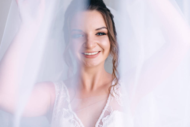 Morgen der Braut. Wunderschöne Braut, schöne Frau mit einem langen Schleier aus Locken in einem weißen Kleid und einem Lächeln. Die Braut vor der Hochzeit. - Foto, Bild