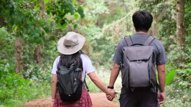 Achteraanzicht van een jong stel wandelaars die hand in hand over de weg wandelen in de natuur. Aziatisch toeristisch paar met rugzakken in het tropisch bos. - Video
