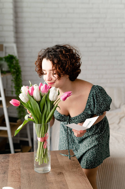 the brunette girl received a bouquet of tulips and a card as a gift. enjoys the smell of flowers in the room - Photo, Image