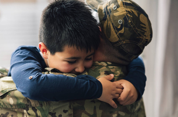 Wojskowy ojciec i jego syn przytulają się w salonie swojego domu. Syn uśmiecha się szczęśliwie do kamery - koncepcja przytulania tatusia -. - Zdjęcie, obraz