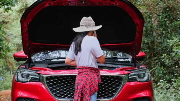 ストレスを受けた女性ドライバーは、オープンフードの前に立ち、スマートフォンを使って車が森の中で故障するのを助けます. - 映像、動画