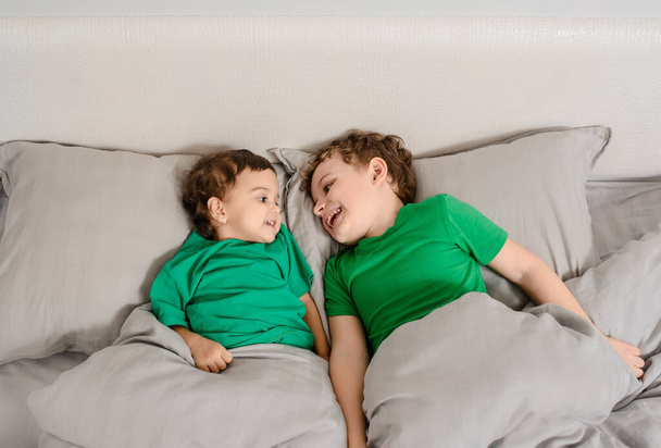Šťastný malý bratříček a sestra v zelených tričkách v posteli se usmívají a smějí. Děti zachrání koncept planety - Fotografie, Obrázek