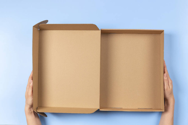 Вид сверху на женские руки, держащие открытую коричневую картонную коробку на светло-голубом фоне. Макет коробки для посылок. Упаковка, покупки, концепция доставки - Фото, изображение