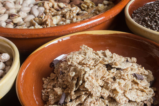Close-up of a pile of roasted rice, whole wheat, and barley flakes with chocolate shavings surrounded by earthenware bowls filled with different dry nuts and legumes. Food and ingredients.	 - Photo, Image