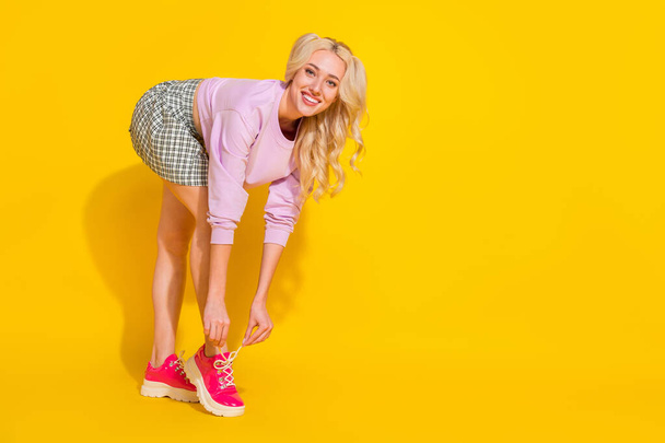 Full length profile portrait of adorable girl tying shoe laces toothy smile isolated on yellow color background - Fotografie, Obrázek