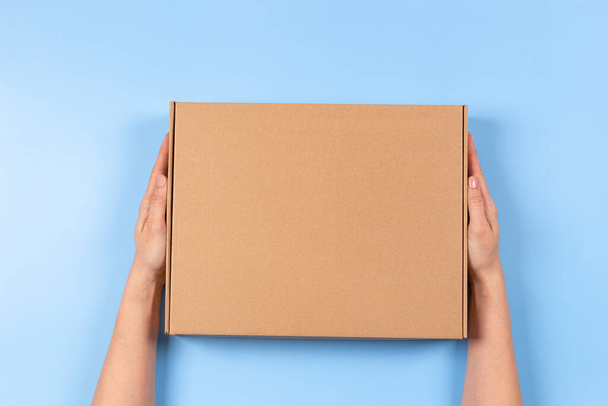 Top view to female hands holding brown cardboard box on light blue background. Mockup parcel box. Packaging, shopping, delivery concept - Photo, Image