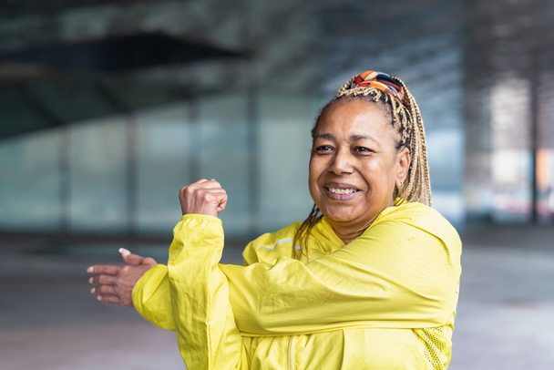 Yaşlı Afrikalı kadın şehirde koşmadan önce esneme hareketleri yapıyor - Yaşlılar Sportif Yaşam tarzı Konsepti - Fotoğraf, Görsel
