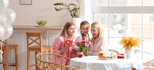 Gelukkige vrouw, haar moeder en dochter met cadeau en bloemen in de keuken. Wenskaart voor Moederdag - Foto, afbeelding